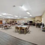 Spacious community room with tables and chairs arranged for group activities, carpeted floor, bookshelves, and wall decorations. The room is brightly lit with large ceiling lights and windows on one side.