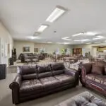 A spacious, well-lit community room with leather sofas in the foreground, dining tables and chairs in the background, and a kitchen area along one wall. The room has beige walls and several windows.