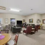 A spacious living area with carpeted floor, featuring sofas, armchairs, a TV, bookshelves, dining tables, and floral decor. Large windows let in natural light, creating a cozy, inviting atmosphere.