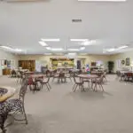 Spacious community room with round tables and chairs, bright lighting, and various decor. There are shelves, cabinets, a counter area at the back, and games or crafts on the side tables. The room has beige carpeting.