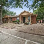 One-story brick duplex homes with beige trim, small front porches, potted plants, and gravel landscaping. Accessible ramps and a shaded area with mature trees are visible. Empty parking spaces are in the foreground.