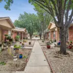 A tidy sidewalk runs between single-story brick homes with small porches, potted plants, and trees providing shade along a well-kept residential courtyard.