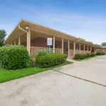 Single-story brick building with a covered porch, beige trim, and neatly trimmed bushes along the front. A sign on the porch reads Leasing Office. Concrete driveway and green lawn surround the building under a clear blue sky.