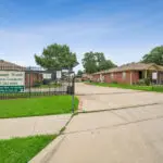 Entrance to Sunny Woods Retirement Community, showing a gated driveway, a sign with contact info, green grass, and several single-story brick buildings on a clear day.
