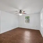 Empty room with white walls, a ceiling fan with lights, hardwood floors, and two windows letting in natural light. An open doorway leads to another part of the home.