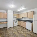 A clean, empty kitchen with light wood cabinets, white appliances including a refrigerator, stove, and dishwasher, black countertops, and a small built-in desk under a window. The floor has a brown tile pattern.