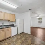 A kitchen with light wood cabinets, a white refrigerator, dishwasher, and double sink, opens to an unfurnished living area with wood floors, white walls, and a ceiling fan near two windows.