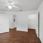 Empty bedroom with white walls, ceiling fan, open white doors, wooden floor, and a walk-in closet with a shelf and hanging rod. The room is well-lit and has a minimal, clean appearance.