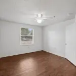 A bright, empty room with white walls, a ceiling fan with a light, a window with blinds, and wood-style flooring. An open door leads to an adjacent room with similar flooring.