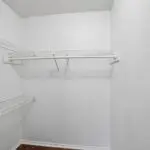 Empty white walk-in closet with wire shelves on the left and back walls, and a hanging rod below the top shelf. The closet has white walls and a brown hardwood floor.