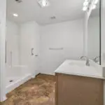 A modern bathroom with a walk-in shower, a wooden vanity with a sink, a large mirror with lights, white walls, tiled flooring, and a towel rack.