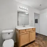 A bathroom with a white toilet, a wooden vanity with a white countertop, a rectangular mirror, and wall-mounted lighting above. The floor has a brown marbled pattern, and a white door is open to the side.