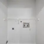 Empty laundry room with white walls, a wire shelf mounted on the wall, hookups for a washer and dryer, and electrical outlets visible below the hookups.