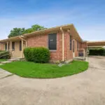 Single-story brick house with tan trim, a covered carport, ramp access, and manicured green grass. The sky is clear and blue, and the home is situated on a concrete driveway in a suburban neighborhood.