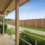 View from a covered porch with a handrail, looking out onto a sidewalk, green grass, bushes, and a tall wooden privacy fence under a clear blue sky.