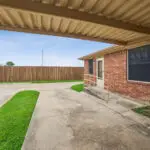 A covered carport with a concrete driveway next to a brick building. The building has a wheelchair-accessible ramp, black window coverings, and an outdoor air conditioning unit. A wooden fence lines the background.