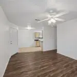 A bright, unfurnished open-plan living area with wood flooring and white walls, leading into a kitchen with light wood cabinets, white appliances, and tile flooring. A ceiling fan with lights is mounted above.