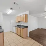 Kitchen with light wood cabinets, white appliances, tile flooring, and adjacent to an open dining/living area with dark wood flooring, white walls, and a ceiling fan near a window and a door.