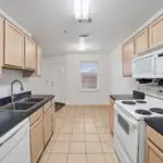 Bright kitchen with light wood cabinets, black countertops, white appliances including a stove and microwave, double sink, dishwasher, and tiled floor. A window and door are visible at the far end of the room.