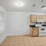 A simple kitchen with light wood cabinets, a white stove, microwave, and tiled floor. There is a large window on the left and white walls throughout the room.