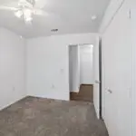 A small, empty bedroom with white walls, a ceiling fan with lights, carpeted floor, a closed closet with white doors, and an open doorway leading to a hallway with wood flooring.