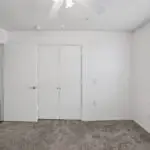 A bright, empty bedroom with white walls, a closed double closet, open door, ceiling fan, large window with blinds, and carpeted floor. Sunlight streams in from the window onto the gray carpet.
