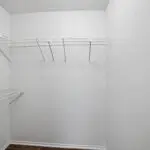 Empty walk-in closet with white wire shelving on two walls, white walls, and a dark wood floor. The closet is well-lit and has a clean, minimalist appearance.