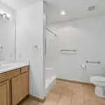 A clean, spacious bathroom with tan tile flooring, a white toilet, a walk-in shower, a large mirror, and a wooden vanity with a white countertop and sink under bright vanity lighting.