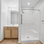 A bathroom with a walk-in shower featuring a grab bar and built-in bench, a towel rack, and a vanity with a sink, mirror, and wooden cabinets under bright lighting.