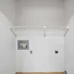 Empty laundry room with white walls, a tiled floor, a single wire shelf, and hookups for a washer and dryer. There are visible water supply valves and electrical outlets on the back wall.