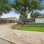 Entrance to Sunny Ridge Retirement Community with a gated driveway, brick buildings, trees, a lawn, and a large sign displaying the community’s name, contact number, and office hours.