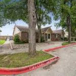 A single-story brick house with a gable roof sits near a curb marked FIRE LANE - NO PARKING. Trees and a small garden area are in front, with a driveway and parking space nearby.