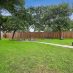 A spacious, grassy yard with large trees, picnic tables, benches, a grill, and a wooden fence surrounding the area. A paved walkway leads through the lawn, and there are potted plants decorating the space.