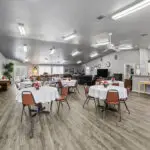 A spacious, well-lit community room with round tables covered in white cloths, brown chairs, a kitchen area, and a lounge section with couches. There are floral centerpieces on the tables and decor on the walls.