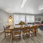 A dining area with a wooden table, eight chairs, a lamp on a side table, floral centerpiece, a red pool table, gray couches, wall art, and large windows with sheer curtains, all under bright fluorescent lights.