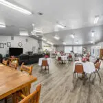 A spacious, well-lit community room with round tables draped in white cloths, each decorated with flowers, brown chairs, a seating area with recliners, wall art, and wood flooring.