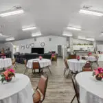 A spacious community room with round tables covered in white cloths, each topped with a small floral arrangement. Brown chairs are arranged around the tables. The room features a kitchen, lounge area, and bright overhead lighting.