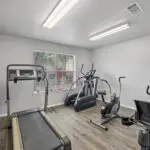 Small fitness room with a treadmill, elliptical machine, stationary bike, and recumbent bike on wood flooring. A window lets in natural light, and overhead lights brighten the clean, gray-walled space.