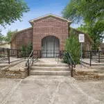 A brick building with a central arched entrance, metal railings, and concrete ramps. A sign on the right reads Sunny Ridge Office. Trees and shrubs surround the building under a blue sky.